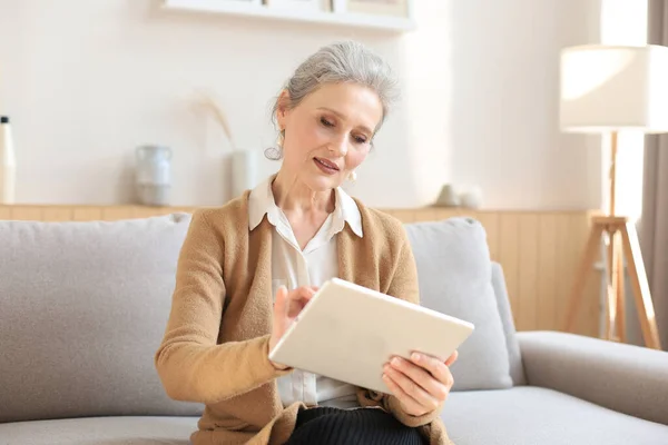 快乐的中年妇女坐在沙发上 使用电脑平板电脑应用程序 观看屏幕 在社交网络上阅读好消息 在网上购物或聊天 — 图库照片