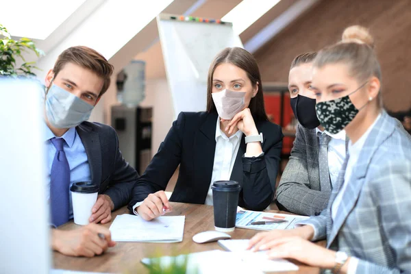일하는 젊은 사업가들의 그룹 사무실 책상에 앉아서 의사 소통을 하는 동안 동료들과 예방 마스크를 쓰고. — 스톡 사진