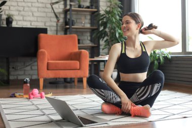 Fitness güzeli ince kadın evde, oturma odasında dizüstü bilgisayarını kullanarak halterler ve bir şişe suyla yerde oturuyor. Evde kal.