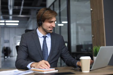Kulaklıklı, gülümseyen bir erkek çağrı merkezi operatörü, modern ofiste oturuyor, bir dizüstü bilgisayardaki çevrimiçi bilgilere danışıyor, müşteriye yardımcı olmak için bir dosyada bilgilere bakıyor.