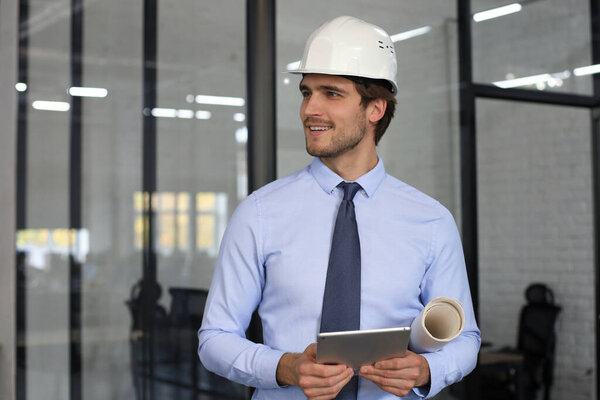 Confident young business man in hardhat holding blueprint and looking away while standing indoors