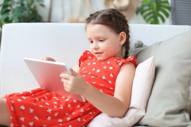 Gülümseyen küçük kız kanepede uzanmış online oyunlar oynuyor, web sörfü bilgileri, tablet üzerindeki komik uygulamaları kullanıyor.