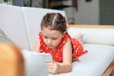 Gülümseyen küçük kız kanepede uzanmış online oyunlar oynuyor, web sörfü bilgileri, tablet üzerindeki komik uygulamaları kullanıyor.