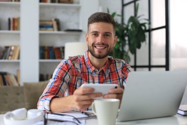 Çekici genç adam evdeki masada oturuyor ve sosyal ağları kontrol etmek için cep telefonunu kullanıyor.