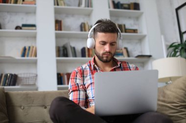 Konsantre olmuş genç serbest çalışan işadamı dizüstü bilgisayarla kanepede oturuyor, uzaktan evde çalışıyor.