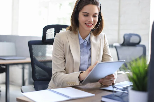 Cheerful female manager sitting at office desk and performing corporate tasks using wireless connection on digital gadgets