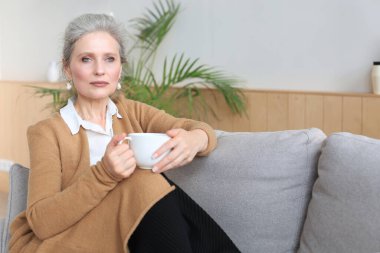 Mutlu olgun bir kadın rahat koltukta dinleniyor kahve ya da çay içiyor, başka tarafa bakıyor, evdeki rahat koltukta dinleniyor sıcak içeceğin tadını çıkarıyor.