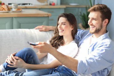 Evdeki kanepede oturup televizyon seyreden ve gülen genç çift.
