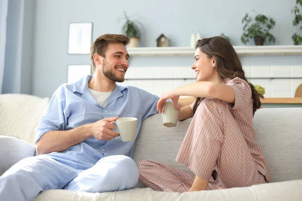 美しい若いカップルでパジャマはお互いを見ていて リビングルームのソファで笑っています — ストック写真