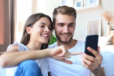 Genç çift evdeki kanepede oturmuş akıllı bir telefonla online içeriği izliyorlar.