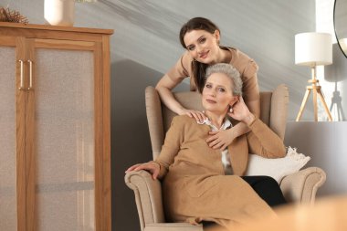 Yaşlı, orta yaşlı bir anne bir sandalyede oturuyor ve kızı sarılıyor, başka tarafa bakıyor, güvenilir akrabaları var.