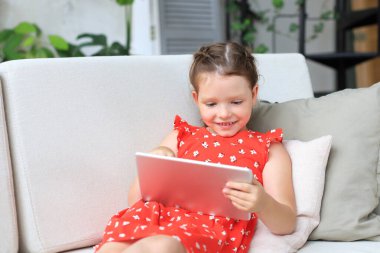 Gülümseyen küçük kız kanepede uzanmış online oyunlar oynuyor, web sörfü bilgileri, tablet üzerindeki komik uygulamaları kullanıyor.