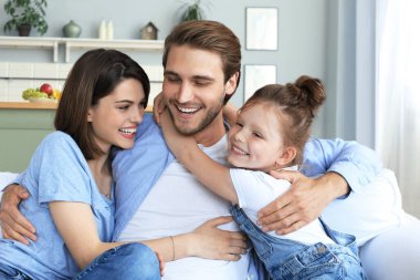 Hafta sonları evde dinlenirken, kanepede birlikte oturan gülümseyen küçük bir kızı olan olumlu, arkadaş canlısı ebeveynler.