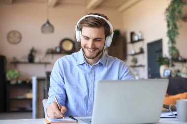 Kulaklıklı kendine güvenen işadamı evde dizüstü bilgisayarla otururken notlar veya mali raporlar yazıyor