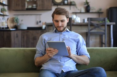 Evde sosyal ağlarla tablet tutan ve kullanan gülümseyen yakışıklı bir adam. İnternetten alışveriş yapan mutlu bir serbest yazar. Oturma odasındaki rahat koltukta oturuyor.