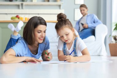 Mutlu anne gülümseyen kız sıcak zeminde uzanıyor yaratıcı aktivitelerin tadını çıkarıyor, albümle resim çiziyor, baba kanepede dinleniyor, aile birlikte boş zaman geçiriyor.