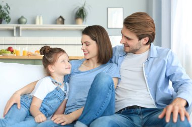 Hafta sonları evde dinlenirken, kanepede birlikte oturan gülümseyen küçük bir kızı olan olumlu, arkadaş canlısı ebeveynler.