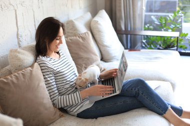 Kanepede dizüstü bilgisayarla oturmuş arkadaşlarıyla sohbet eden küçük bir köpek gibi gülümseyen genç bir kadın.