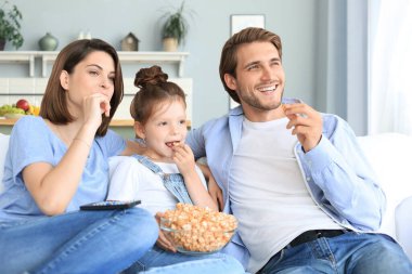 Mutlu ve çocuklu bir aile kanepede oturur televizyon izler ve patlamış mısır yer, genç ebeveynler kızlarını kucaklar birlikte kanepede dinlenirler..