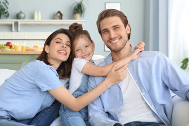 Hafta sonları evde dinlenirken, kanepede birlikte oturan gülümseyen küçük bir kızı olan olumlu, arkadaş canlısı ebeveynler..