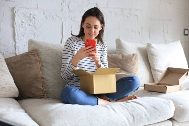 Güzel genç bir kadın elinde karton bir kutu tutuyor ve evdeki kanepede oturan akıllı telefonu açıyor.