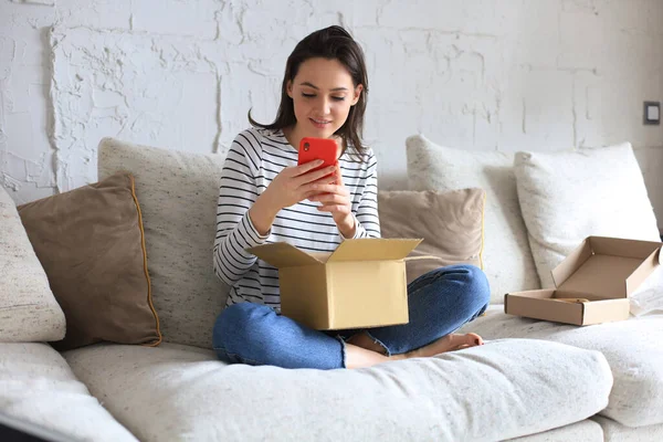 美しい若い女性は段ボール箱を持っていて 家でソファに座っているスマートフォンをアンパックしています — ストック写真