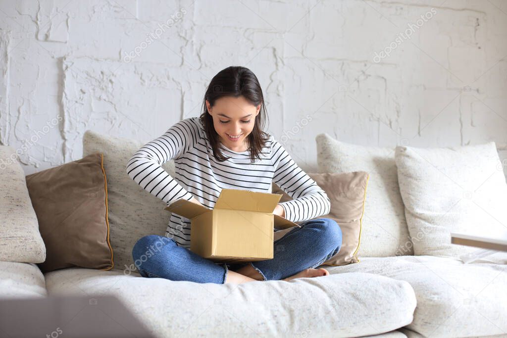 Beautiful young woman is holding cardboard box and unpacking it sitting on sofa at home