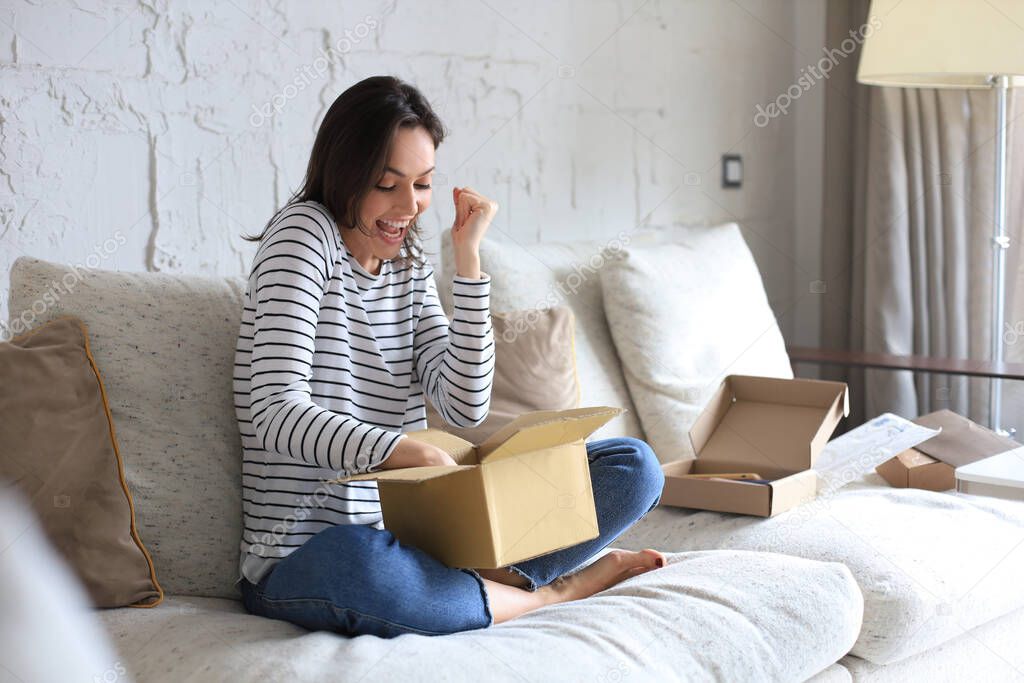 Pretty woman is unpacking parcel, expressing joy, sitting on sofa at home