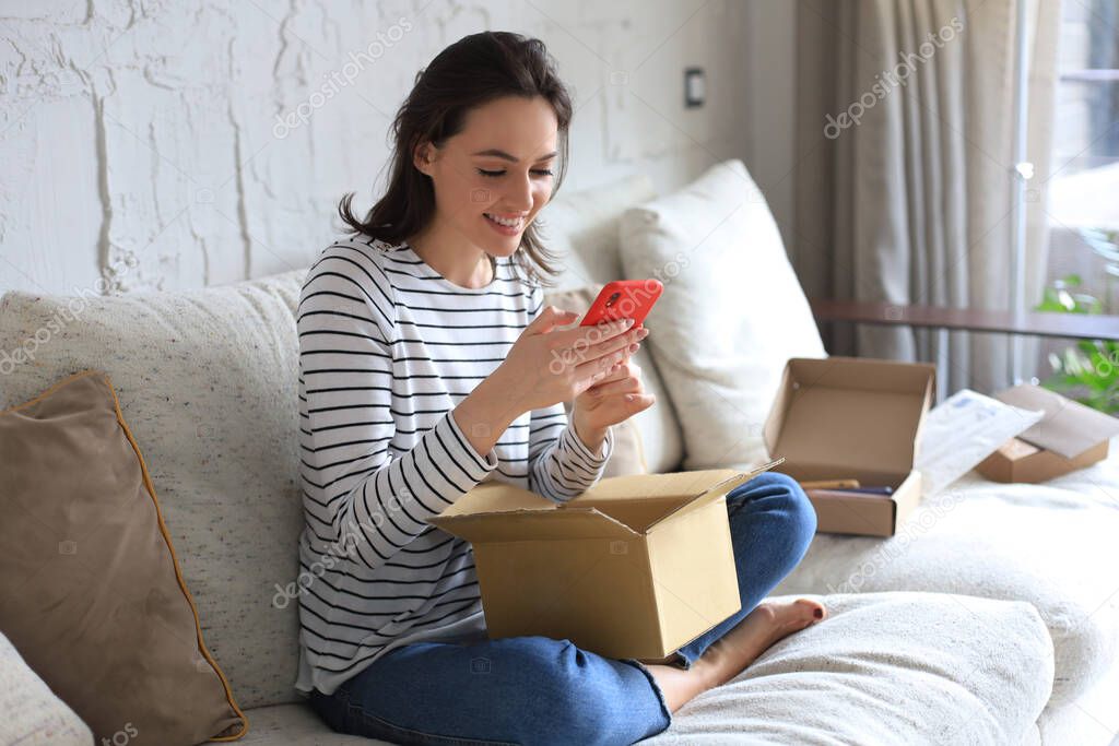 Beautiful young woman is holding cardboard box and unpacking smartphone sitting on sofa at home
