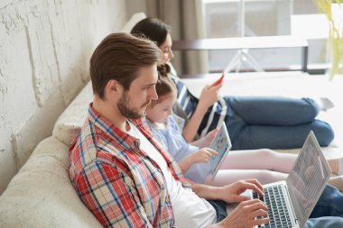 Baba, anne ve kız oturma odasındaki koltukta oturan elektronik aletleri kullanıyorlar.