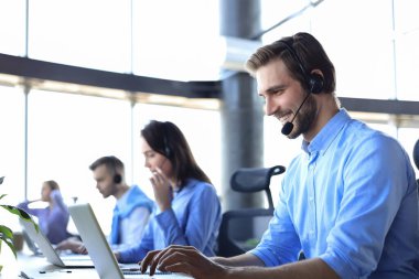 Kulaklıklı, gülümseyen erkek çağrı merkezi operatörü, modern ofiste arka planda meslektaşlarıyla oturuyor, online danışmanlık yapıyor.
