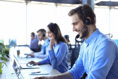 Kulaklıklı, gülümseyen erkek çağrı merkezi operatörü, modern ofiste arka planda meslektaşlarıyla oturuyor, online danışmanlık yapıyor.