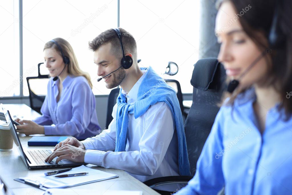 Smiling male call-center operator with headphones sitting at modern office with collegues on the backgroung, consulting online