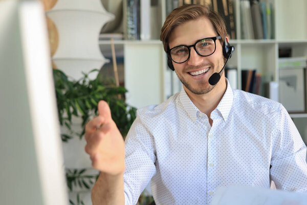 Cheerful young support phone male operator in headset, at workplace while using computer, help service and client consulting call center concept.