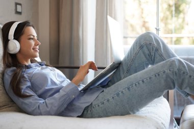 Güzel kız evdeki kanepede dizüstü bilgisayar kullanıyor. Müzik dinlemek, zor bir haftadan sonra rahatlamak.