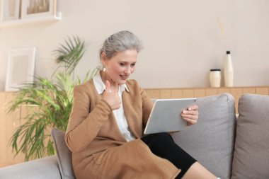 Neşeli orta yaşlı kadın kanepeye oturuyor, bilgisayar uygulamaları kullanıyor, ekrana bakıyor, sosyal ağda iyi haberler okuyor, alışveriş yapıyor ya da internette sohbet ediyor.