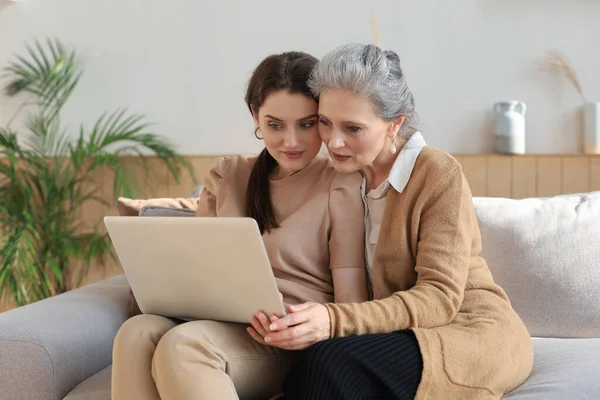 Happy elderly middle mother sitting on couch with her daughter, looking at laptop. Young woman showing video, photos to mommy, trusted relations. Family concept