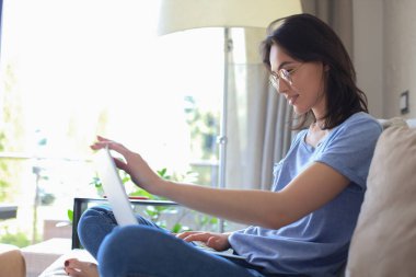 Kanepede dizüstü bilgisayarla oturan ve arkadaşlarıyla sohbet eden gülümseyen genç bir kadın.