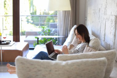 Kanepede dizüstü bilgisayarla oturmuş arkadaşlarıyla sohbet eden küçük bir köpek gibi gülümseyen genç bir kadın.