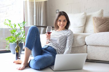 Gülümseyen genç bir kadın dizüstü bilgisayarla yerde oturuyor arkadaşlarıyla sohbet ediyor, şarap içiyor.