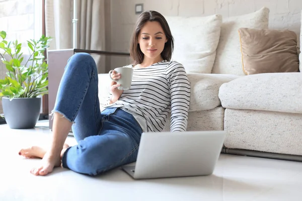 自宅で床に座ってコーヒーを飲みながらノートパソコンで働く可愛い女性 ストック写真