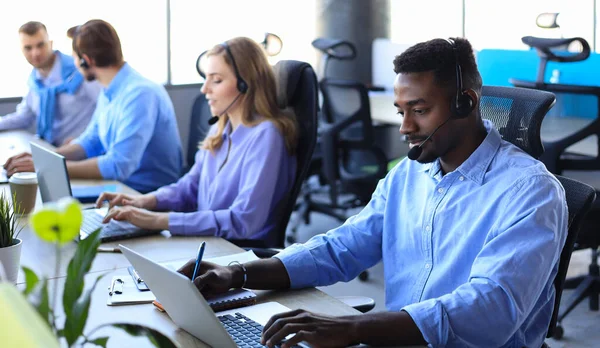 Mosolygó afrikai férfi call-center üzemeltető fülhallgató ül a modern irodában kollégák a háttérben, tanácsadás online. — Stock Fotó