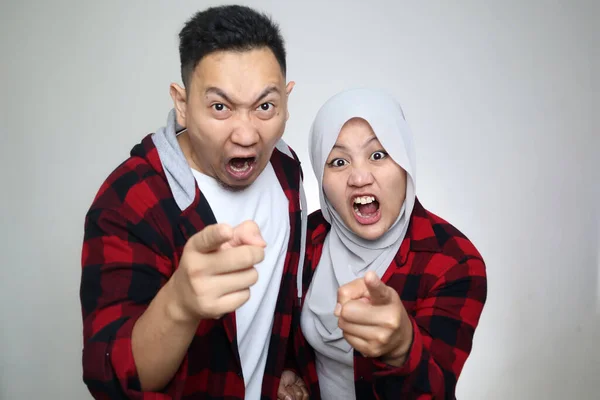Angry Asian muslim couple, husband and wife screaming and pointing to camera, aggressive furious expression, over grey background