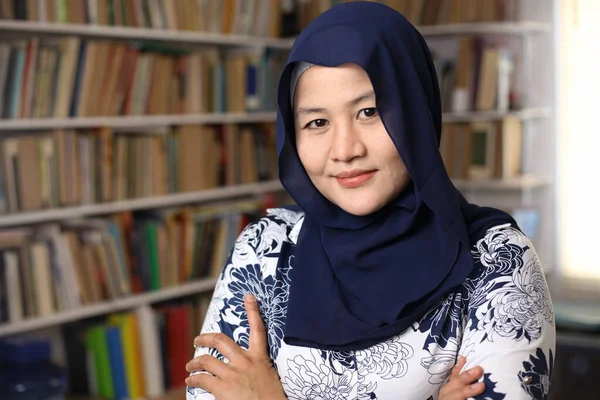 陽気なアジア系ムスリム女性図書館員のヒジャーブ姿の肖像 カメラを見て笑顔が図書館の本の殻に対する腕の自信のジェスチャーを交差 — ストック写真