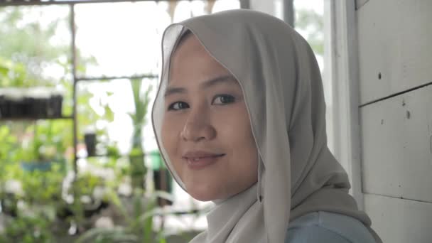 Asiática musulmana mujer usando hijab sonriendo a cámara, mujer stiting relajarse fuera de su casa — Vídeos de Stock