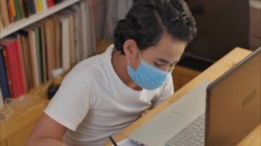 Koruyucu maske takan Asyalı bir öğrenci kütüphanede dizüstü bilgisayarla çalışarak öğreniyor, evden okula geçiyor.