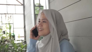 Mutlu Asyalı Müslüman kadın evinin önünde telefonla konuşuyor.