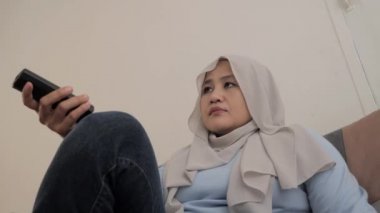 Mutlu Asyalı Müslüman kadın elinde kumandayla televizyon izlerken gülümsüyor.