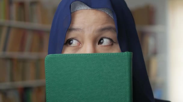 Портрет азиатской мусульманки, закрывающей лицо книгой в библиотеке, думающей жестом — стоковое видео