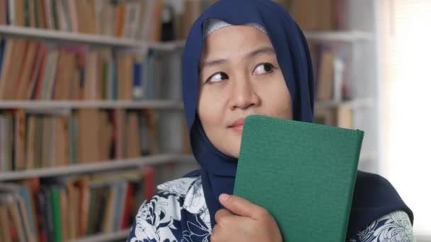 Portrait of Asian muslim woman holding book in library, thinking gesture and smile to camera — Stok Video